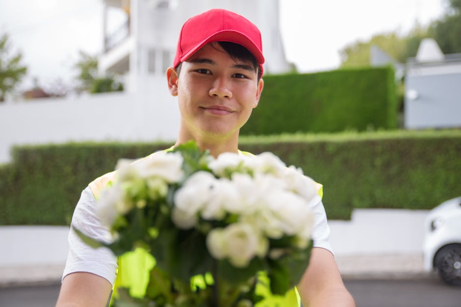 A young man wearing a red cap and a high-visibility vest is standing outdoors on a street, holding a bouquet of white flowers with green foliage. The bouquet appears freshly arranged with delicate pet