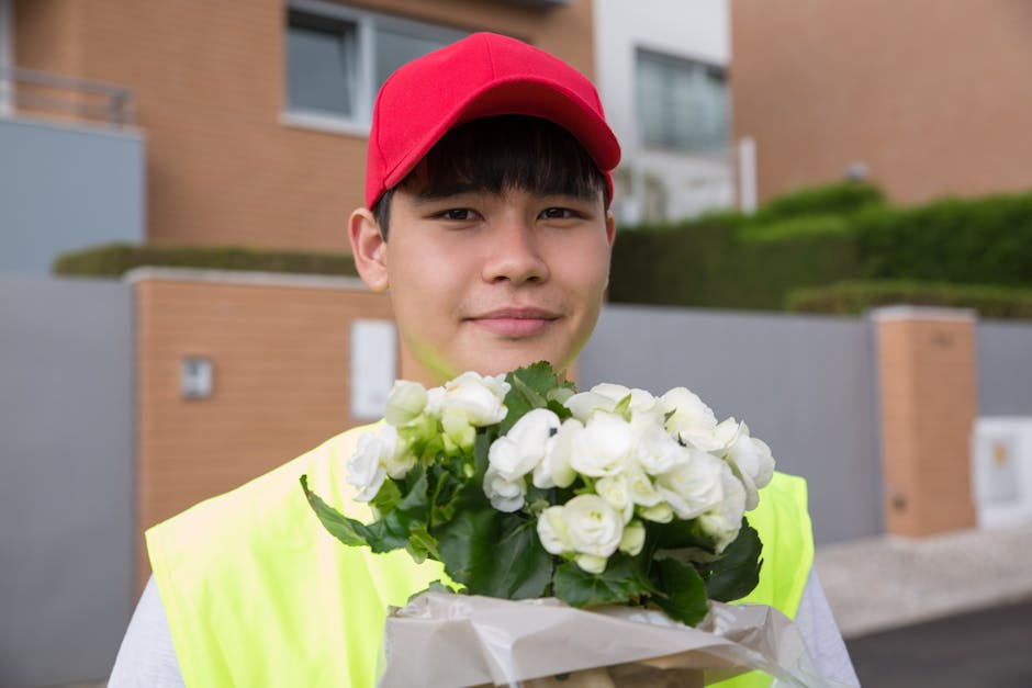 A young male flower delivery courier wearing a red cap and high-visibility yellow vest holds a bouquet of fresh white roses wrapped in white paper, standing outdoors in front of modern residential bui