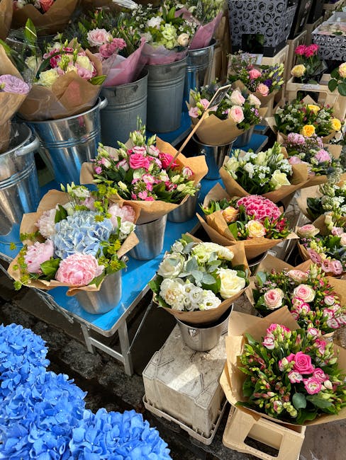 An outdoor display of fresh floral bouquets arranged in metal buckets, featuring a variety of pastel-colored roses, hydrangeas, and carnations, wrapped in brown craft paper and pink tissue paper. The 