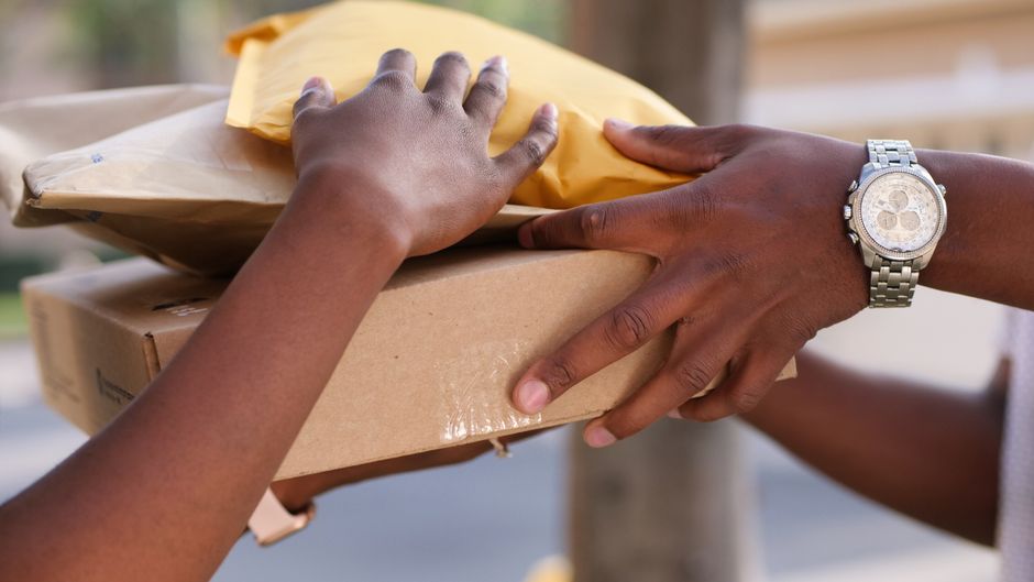 Close-up of two hands handling a package for flower delivery, one hand gripping a brown cardboard box and the other adjusting the yellow wrapping paper on a floral gift. The person on the right is wea