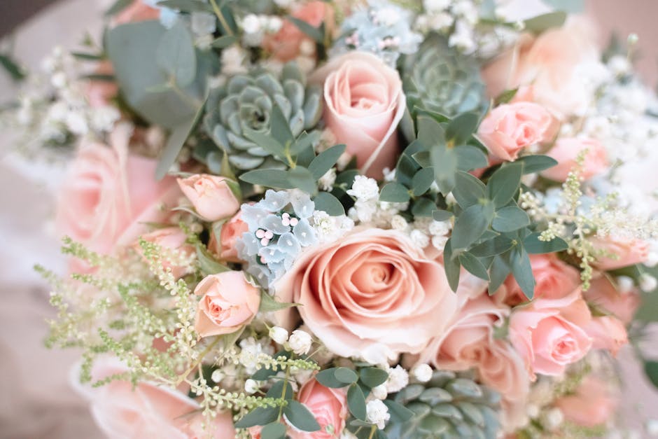A close-up view of a floral arrangement featuring large, soft pink roses and smaller pink spray roses, complemented by silvery-green eucalyptus leaves and delicate white baby's breath. The flowers are