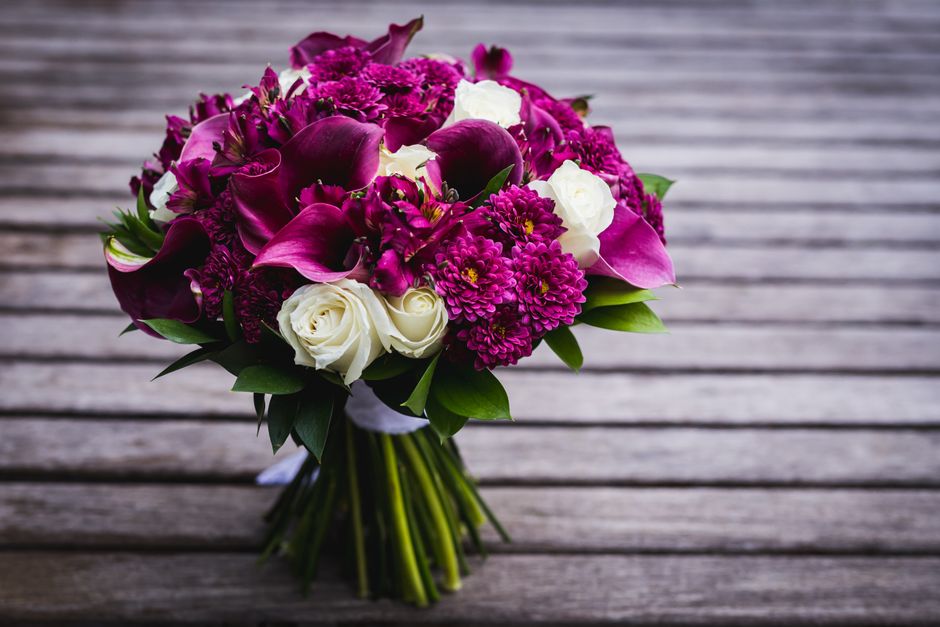 A bouquet arrangement featuring deep magenta calla lilies, vibrant pink chrysanthemums, and clusters of small magenta flowers, accented with white roses. The flowers are wrapped in elegant white tissu