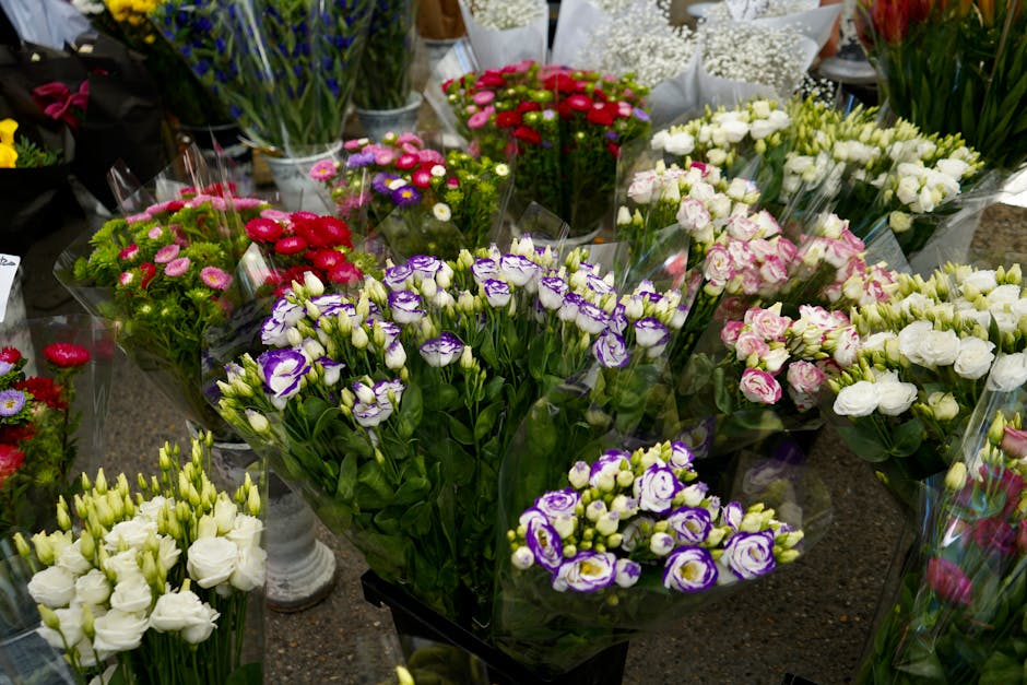 A variety of fresh floral arrangements displayed on a retail table, featuring bouquets wrapped in clear cellophane. The flowers include purple and white lisianthus, pink and white roses, small red and