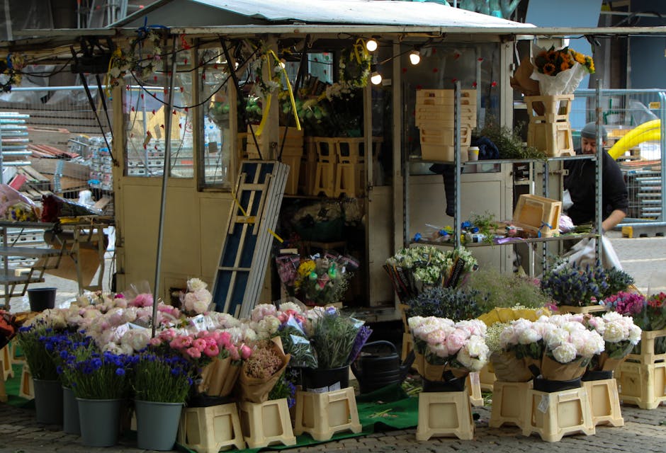 A street-level flower stall displaying a colorful array of fresh flowers and floral arrangements in various shades of pink, purple, blue, and white. The flowers are wrapped in brown paper and arranged