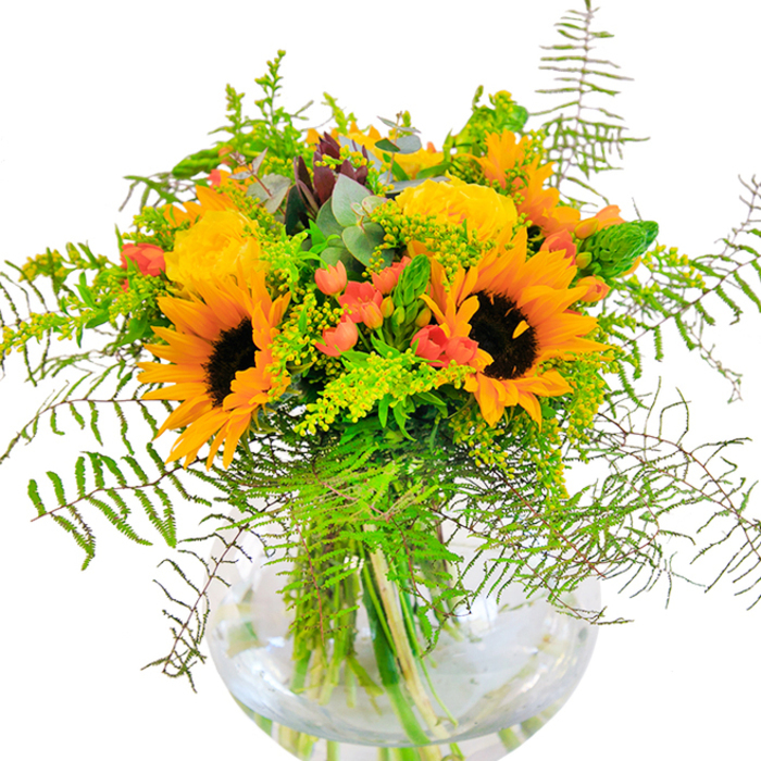 On first sight, this stunning bouquet is pure sunlight captured in blooms-a composition that seems to glow with the untamed brightness of Rotherhithe's open spaces. Prominent sunflowers take centre stage, their rich brown centers bordered by lemon and amber petals, unfurling toward you like a promise of happiness. Nestled intimately beside them, garden roses in saturated yellows, their tightly coiled petals soft as silk, add romance and structure that echoes the timeless gardens found near Canada Water. Vivid red Hypericum berries sparkle among the foliage, glossy and jewel-like, standing out boldly against a backdrop of deep and minty eucalyptus greens. The arrangement tumbles gracefully out of a crystal-clear, rounded vase-its form reminiscent of the tranquil curves of Greenland Dock-and reveals a tangle of strong green stems, alive and thriving in pure, clear water. Texture reigns here: feathery fern fronds arch delicately, while light sprigs of goldenrod and airy greenery offer dimension and movement. There's a palpable freshness, as if every element has just been gathered from the riverside on a dewy summer morning. In the softly-lit room, the bouquet casts golden shadows, radiating warmth and jubilation. Perfect for marking milestones-anniversaries, housewarmings, or family gatherings within Rotherhithe-this arrangement presents a harmonious blend of joy and sophistication, embodying the vibrant character and natural splendor of its local roots.